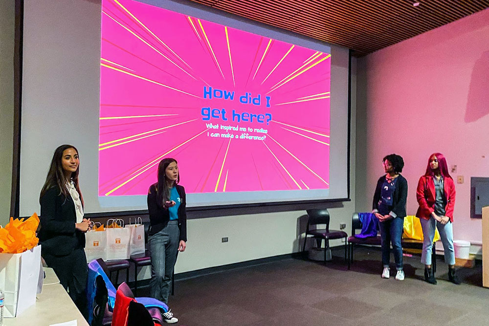 Four women speak to a crowd with a screen reading "How did I get here?" behind them.